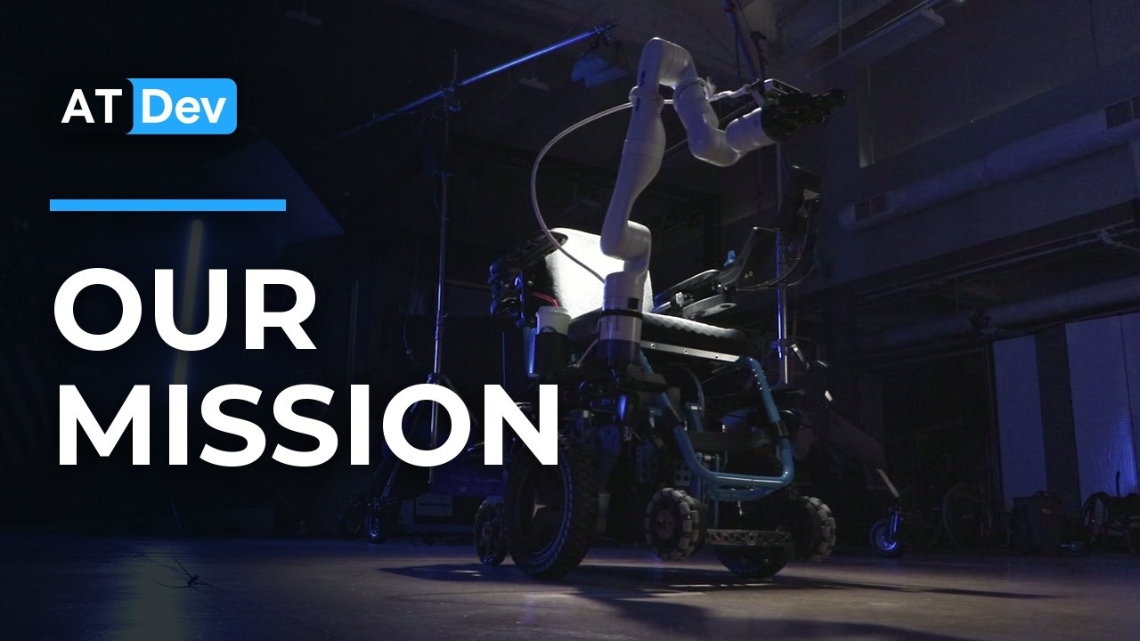 A robotic wheelchair prototype under studio lights in a dark workshop. A white robotic arm is mounted behind the seat and reaches up over it, the frame rides on knobbly all-terrain wheels, and the words OUR MISSION are set large across the left of the picture.